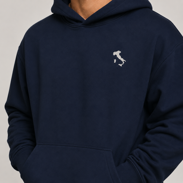 A close-up of a person wearing a navy blue hoodie with a small white embroidered map of Italy on the left chest, set against a plain light gray background.